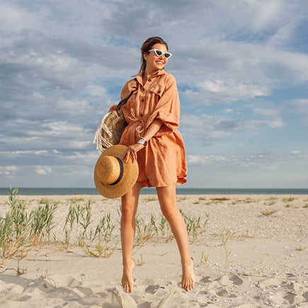Woman in a peach dress standing on a sandy beach with a straw hat and bag.